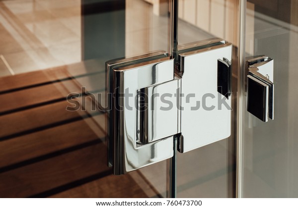 Close metal silver hinge is mounted on\
the glass construction of the door to the sauna, bathroom or shower\
cubicle against the background of the wooden\
floor