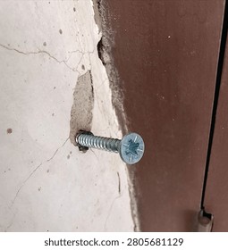 Close up of a metallic self-tapping screw driven into a cracked concrete wall next to a brown metal frame