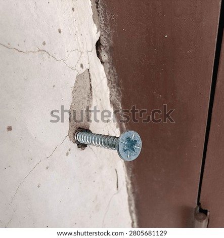 Close up of a metallic self-tapping screw driven into a cracked concrete wall next to a brown metal frame
