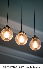 Close Up Mid Century Modern Remodeled Home Dining Living Room Ceiling Chandelier With Three Hanging Incandescent Illuminated Light Bulb In Glass Globe Orbs On Black Electrical Cable With Crown Molding