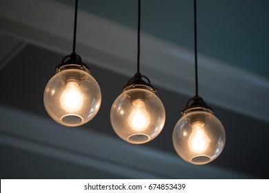 Close Up Midcentury Modern Remodeled Home Dining Living Room Ceiling Chandelier With Three Hanging Incandescent Illuminated Light Bulbs In Glass Globe Orbs On Black Electrical Cable With Crown Molding