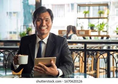 Close Up Middle Age Businessman In Formal Suit Sitting And Holding A Coffee Cup With Tablet, Looking At Camera With Milk Froth On His Mouth At Coffee Shop.