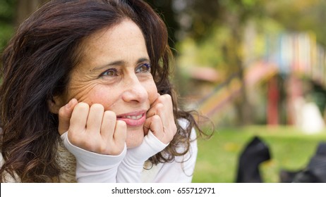 Close up of middle aged lovely brunette lady with hands on face with hopeful look in her eyes while resting in the park. Pretty mature woman, entrepreneur, positive thinking concepts