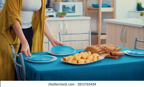 Close Up Of A Middle Aged Woman And Older Senior Have Fun Working Together Setting The Dinner Table In Kitchen, While Men Talking In Background During A Relaxing Family Day.