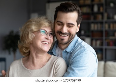 Close up middle-aged mother and her grown up millennial son embracing standing in living room at home, intergenerational family laughing enjoy time together. Care and love, best closest friend concept