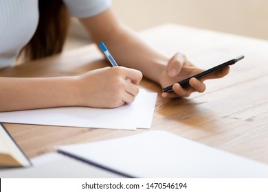 Close Up Millennial Female Student Sitting At Desk, Holding Phone In Hand, Checking Important Information Or Data, Taking Down Notes On Paper. Focused Young Girl Preparing For College Test Or Exam.