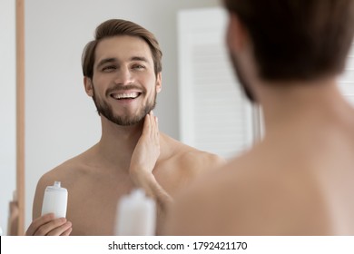 Close up mirror reflection overjoyed young man applying moisturizing aftershave lotion, standing in bathroom, happy smiling handsome guy enjoying skincare routine procedure after shaving