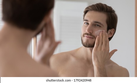 Close up mirror reflection satisfied young man applying face moisturizing cream or lotion on cheek, handsome bearded male enjoying skincare procedure, standing in bathroom after shower