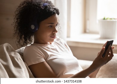 Close Up Mixed Race Woman Wearing Headphones Holding Smart Phone Listen Music, African 30s Female Resting Leaned On Sofa At Home Search Tune Choose Playlist Enjoy Song Using Online Application Concept