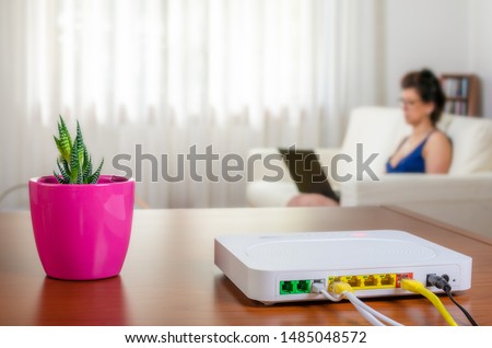 Close up of a modem router on a table in a living room with a woman using a laptop while sitting on the sofa in background