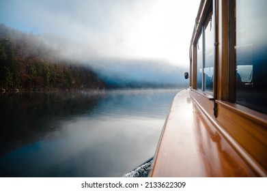 Close up of modern wooden boat floating on lake Konigsee in Berchtesgaden. Winter fog over forest and mountains with reflection on water surface.