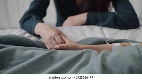 Close up of mother holding hand of sick child having dropper and lying in bed at hospital. Cropped shot of sick kid resting in bed at ward and parent visiting and supporting at bedside - Powered by Shutterstock