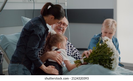 Close Up Of Mother And Kid Surprising Grandpa With Visit In Hospital Ward, Bringing Flowers To Aged Man With Sickness. Family Visiting Senior Patient In Bed, Giving Support. Handheld Shot