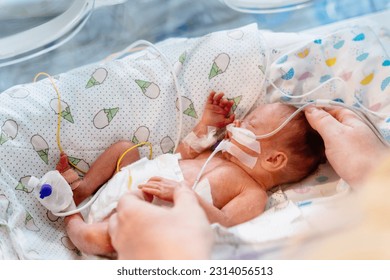 Close up of mother's hands holding new born baby born at 32 weeks gestation in intensive care unit in a medical incubator.