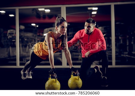 Close up of motivated focused attractive young woman doing push ups exercise on the kettlebells in the gym with her handsome muscular personal trainer.