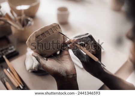 Close up of a multicultural pottery class student's hands decorating clay work and earthenware with pottery tool. Cropped picture of an african american craftswoman's hands decorating pottery on class
