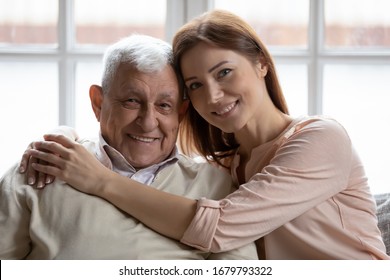Close up of multi-generational relatives people family portrait concept. Loving caring grown up granddaughter hugs elderly 75s grandfather seated on sofa look at camera and smile capture lovely moment