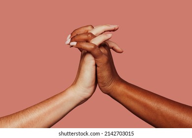 Close up multiracial woman couple with black and caucasian hands holding each other wrist in tolerance unity love and anti racism concept. Pink background.