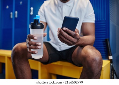 Close up of muscular black sportsman using smartphone in locker room and holding water bottle