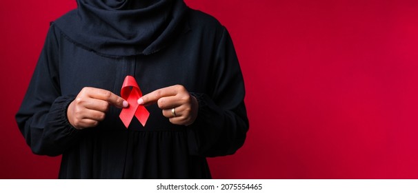 Close up of muslim asian woman in black dress holding and showing red awareness ribbon in her hands on red background