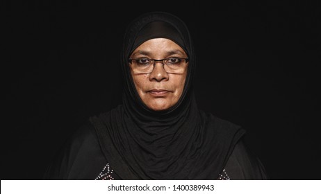 Close up of a muslim woman in hijab isolated on black background. Arabic woman wearing eyeglasses and a black hijab looking at camera.