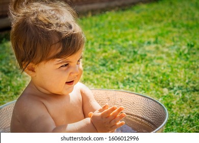 Close Up Of Naked Infant Baby Girl Sitting In A Tin Ice Bucket Full Of Water Clapping Outdoors On Green Grass Background