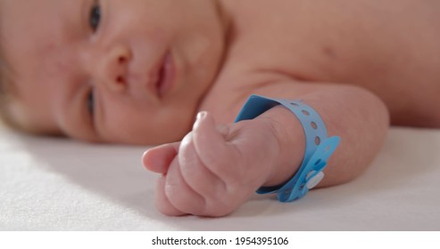 Close Up Of Newborn Baby With Nametag Bracelet. Infant Lying In Crib In Hospital. New Born Baby First Days Of Life In Delivery Room. Healthcare Concept