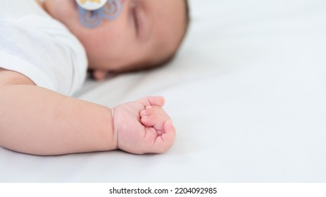 Close Up Of Newborn Boy Hand On White Sheet Background. Baby Sleeping On The Bed. A Beautiful Conceptual Image Of Childhood. Family And Home Concept With Copy Space.