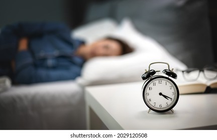 Close Up Of Night Clock Standing On White Table Near Bed. Caucasian Woman In Pajamas Sleeping Peacefully On Blur Background. Sweat Dreams At Bedroom.