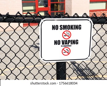 Close Up Of A No Smoking, No Vaping Sign Fixed On Metal Chain Fence In Front Of School Playground And Entry Area. Smoke-Free Act Prohibits The Smoking Of Tobacco, The Vape Of Electronic-cigarettes.