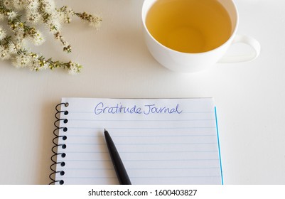 Close up of notebook and pen with handwritten Gratitude Journal text on table with cup of tea and flowers (selective focus)