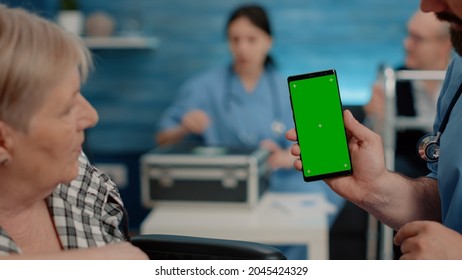 Close Up Of Nurse Vertically Showing Phone With Green Screen To Senior Patient Living In Nursing Home. Medical Specialist Holding Smartphone With Chroma Key And Isolated Mockup Template