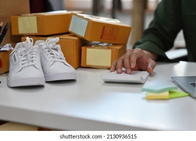 Close Up An Office Man Sitting In The Office Using A Laptop And A Calculator, Boxes Piling Up On The Table And White Sneakers On The Side, For SME,start Up, Financial, E-commerce And Delivery Concept.