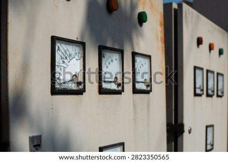 Close up of old electrical control panel boxes with analog gauges, meters, switches, and colorful indicator lights on a wall. Vintage industrial power system or generator control dashboard.