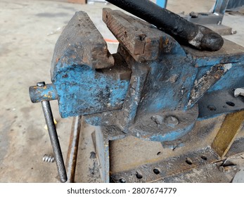 Close up of an old rusty blue bench vice gripping a metal rod in a grunge garage setting