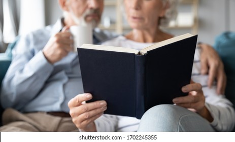 Close Up Older Couple Reading Book Together At Home, Mature Woman Holding Book, Reading Aloud While Man Hugging Her And Drinking Tea Or Coffee, Spouses Enjoying Literature, Common Hobby