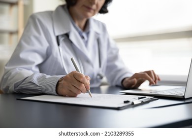 Close Up Older Female General Practitioner Doctor Handwriting Notes On Clipboard, Web Surfing Information Online On Computer, Managing Patient's Appointment Or Writing Healthcare Prescriptions.