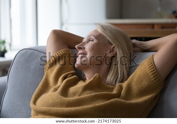 Close up older peaceful woman put hands behind\
head rest leaned on sofa cushions smile looks into distance, take\
break at home, enjoy fresh conditioned air inside. Hotel\
accommodation, relax\
concept