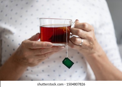 Close Up Older Woman Holding Glass Cup Of Black Tea With Tea Bag Inside In Hands With Manicure, Mature Female Relaxing At Home, Drinking Enjoying Hot Beverage In Mug In The Morning