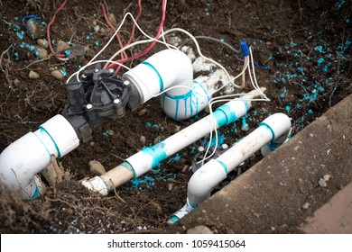 Close Up On An Automated Underground Irrigation Sprinkler System, With Electric Solenoid Valves Beside Water Pipes And Buried In Dirt