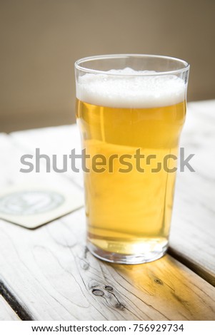Close up on a backlit beer glass filled with a pale blonde ale, on a wood picnic table with a coaster in the background, at a local craft brewery