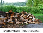 A close up on a big stack of chopped wood having the form of quarter and semi logs gathered next to a vast river or lake covered with reeds with a small axe still being attached to one of the logs