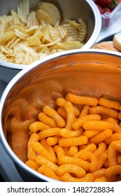 Close up on a bowl of puffed cheese snacks beside potato chips, in a party food background