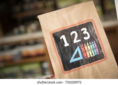 Close Up On A Brown Paper Lunch Bag, With A Hand Painted Educational Graphic For Learning Math With Numbers And Geometry Triangle, Beside Coloring Crayons