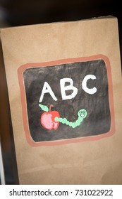 Close Up On A Brown Paper Lunch Bag,  With A Hand Painted Educational Graphic For Learning The Alphabet With And Apple Book Worm