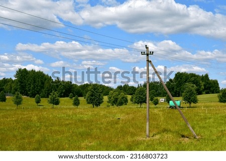 A close up on a concrete power pylon located in the middle of a field, meadow, or pasturelant, surrounded with shrubs, farming equipment and other items seen on a cloudy summer day