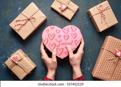 Nahaufnahme von weiblichen Händen, die ein Geschenk in einem rosafarbenen Herz Geschenke für Valentinstag, Geburtstag, Muttertag halten. Flat lay. Symbol der Liebe. Valentinstag Hintergrund mit Geschenkbox auf Beton-Brett.