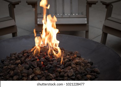 Close Up On Glowing Lava Rock Coals In A Black Iron Fire Pit, With Motion Blurred Flames, And Chairs In The Dark Background