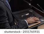 Close up on hand of African American male politician resting on book, while official swearing to obey law standing at podium on stage or in court