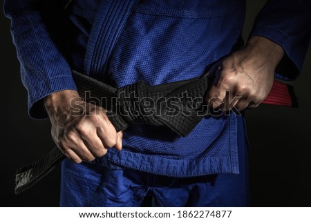 Close up on hand of unknown caucasian man holding brazilian jiu jitsu bjj black belt around his waist while wearing kimono gi in dark - martial arts mastery skill and confidence achievement concept